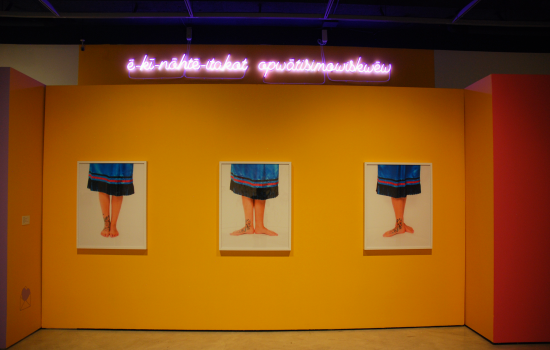 A yellow wall with 3 canvas images of feet painted with floral designs and a neon pink sign above