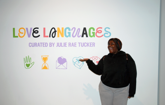 A student stands in front of a colourful sign on a white wall that says "Love Languages" curated by Julie Rae Tucker