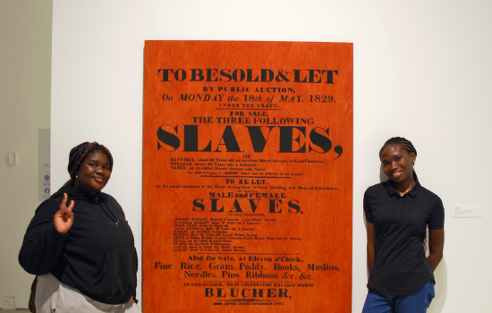 2 students stand on either side of a piece of artwork depicting a bill for the sale of slaves