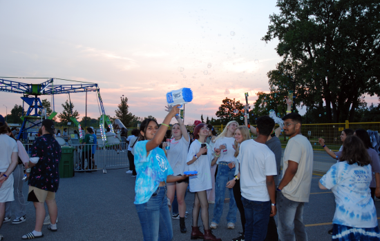 A crowd of students inside the colour party. One is wearing a tie dye shirt and holding a bubble machine. 