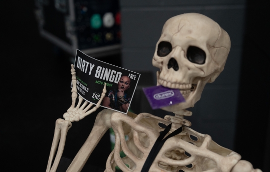 A plastic skeleton with a condom in its mouth and holding a flyer for Dirty Bingo